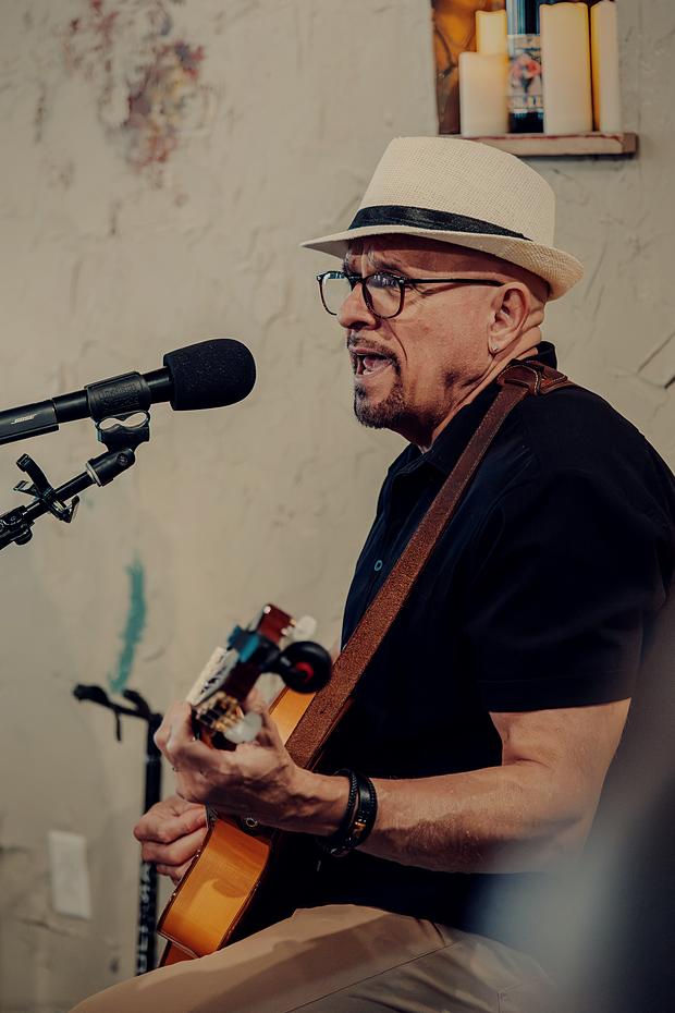 A singer at the microphone in a panama hat, guitar strap over his shoulder.